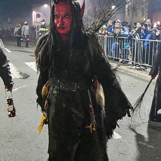 Eine Person in einem rotgesichtigen Kostüm mit Hörnern geht nachts auf einer Straße. Mehrere Leute stehen hinter einem Metallzaun und schauen zu.