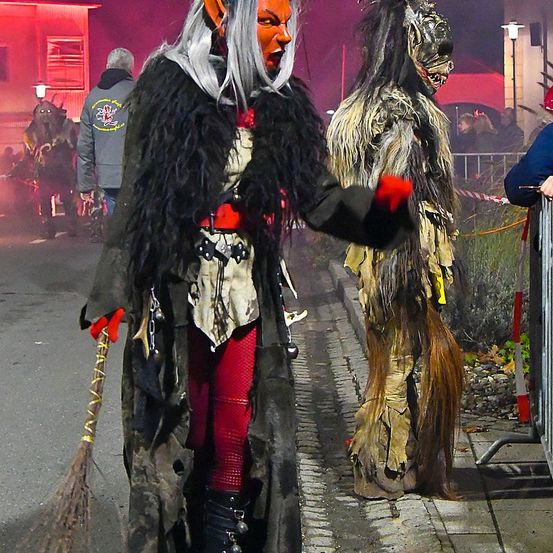 Zwei Menschen in gruseligen Kostümen stehen auf dem Bürgersteig. Einer trägt ein Hexenkostüm mit Besen und der andere ist als gruseliges Monster verkleidet. Im Hintergrund sind Menschen, die sie beobachten.