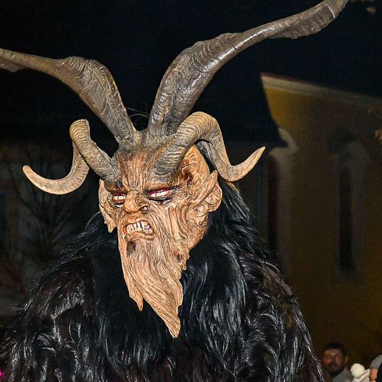 Eine Person in einer Hörnermaske mit einem gruseligen Gesicht steht. Die Person trägt einen Pelzmantel. Hinter ihr stehen ein paar Leute. Das Licht fällt auf die Person.