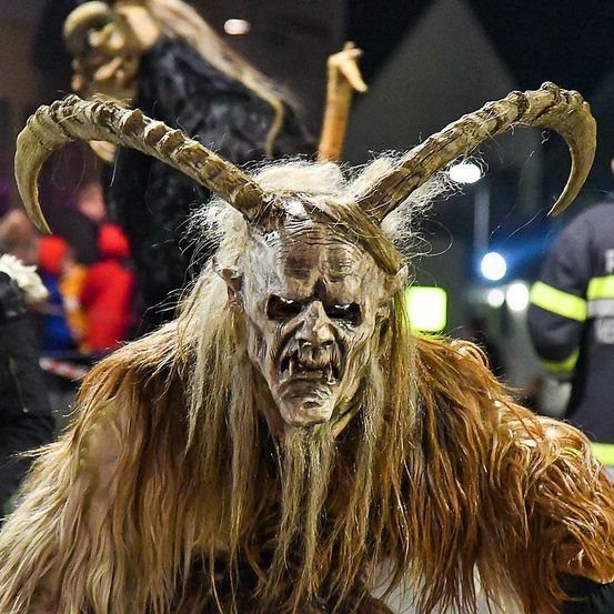 Eine Person in einem pelzigen Kostüm mit Hörnern steht bei Nacht vor einer Menge. Im Hintergrund ist eine Person in einem schwarzen Kleid zu sehen.