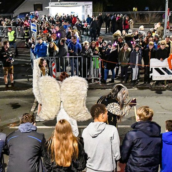 Eine große Menschenmenge steht hinter Absperrungen und beobachtet eine Veranstaltung. Einige tragen Kostüme, eine Person mit Engelsflügeln und eine andere mit einer Maske. Die Atmosphäre ist festlich, mit einem Gebäude und Straßenlaternen im Hintergrund.