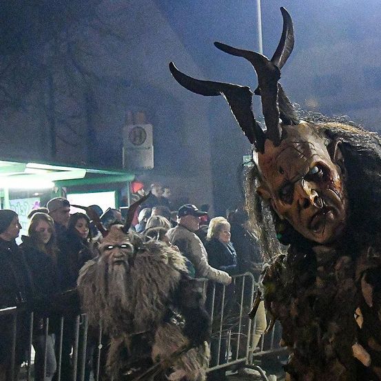Zwei Personen in Monsterkostümen, eine mit Hörnern und einer Maske, stehen vor einer Menschenmenge. Sie sind von einem Metallzaun umgeben. Im Hintergrund ist ein Schild zu sehen.