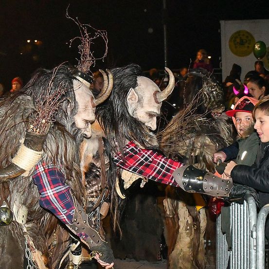 Zwei Personen in Hörnermasken interagieren mit einem Kind während eines Nachtfestivals. Eine Menschenmenge steht hinter einem Metallzaun und beobachtet.