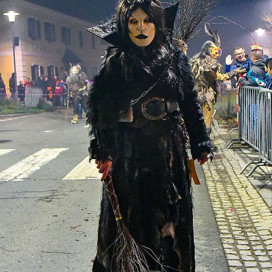 Eine Frau als Hexe verkleidet mit einem Besen geht nachts die Straße entlang. Hinter ihr sehen die Leute eine Parade mit anderen kostümierten Charakteren. Die Straße ist mit Absperrungen und einem Gebäude mit Fenstern gesäumt.