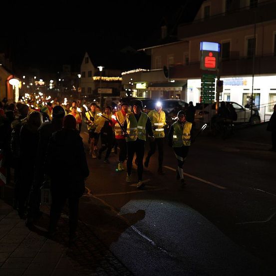Ein nächtliches Straßenereignis zeigt Menschen, die mit Stirnlampen und Fackeln laufen, zusammen mit Zuschauern auf dem Bürgersteig. Gebäude mit Lichtern und Schildern sind sichtbar.