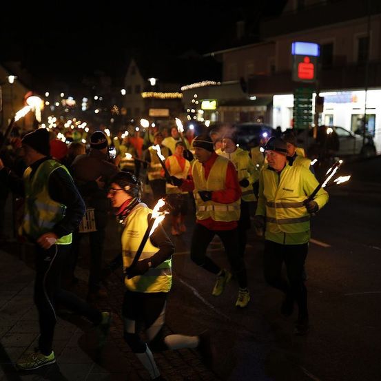 Eine Gruppe von Läufern in reflektierenden Westen, die brennende Fackeln halten, läuft nachts auf einer Straße. Im Hintergrund sind Gebäude mit Lichtern und einem blauen Schild zu sehen.