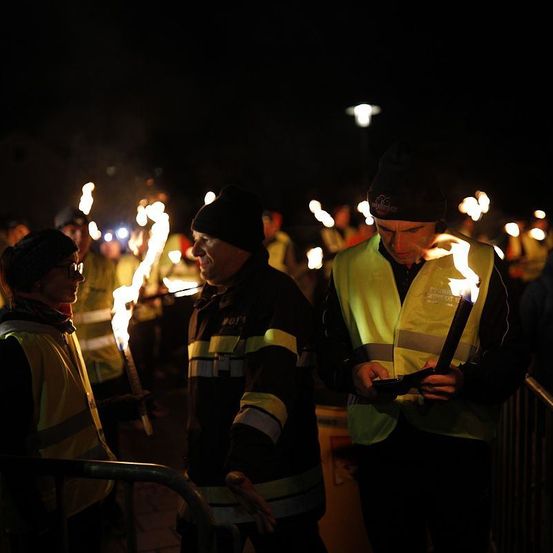 Drei Personen in reflektierenden Westen und Mützen stehen zusammen und halten brennende Fackeln. Sie scheinen Teil einer größeren Gruppe zu sein, mit weiteren Menschen und Fackeln im Hintergrund sichtbar.