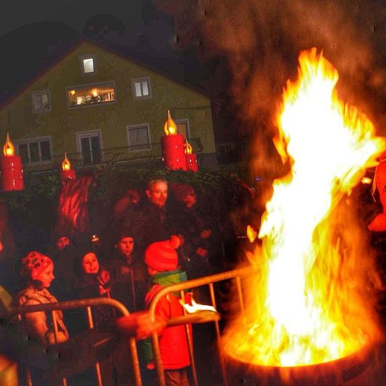 In der Nacht versammeln sich mehrere Personen um ein großes Feuer, möglicherweise für eine Feier. Einige tragen Mützen und Jacken. Rote Kerzen sind um das Feuer herum aufgestellt. Ein Haus mit leuchtenden Fenstern ist im Hintergrund.