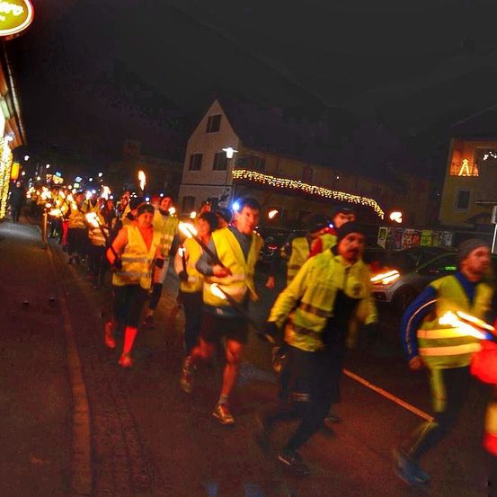 Eine Gruppe von Menschen in reflektierenden Westen läuft bei Nacht mit brennenden Fackeln die Straße entlang. Die Straße ist nass, und Gebäude säumen die Straße.