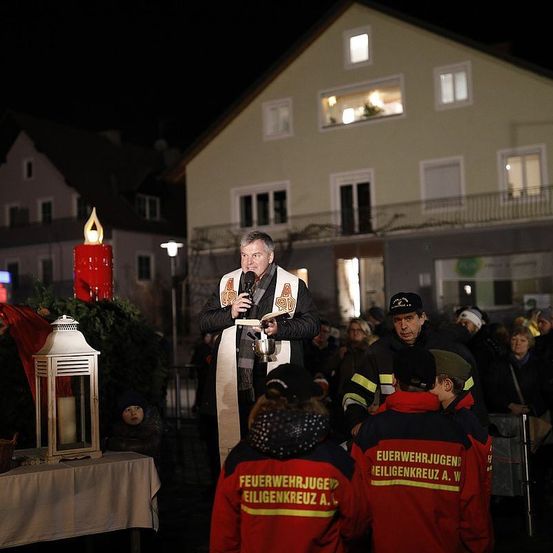 Ein Priester spricht eine Menschenmenge bei Nacht an, mit einer brennenden Kerze auf einem Tisch, zwei Feuerwehrleuten in Uniform und einem Gebäude im Hintergrund.