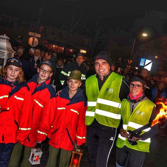 Mehrere Menschen in roten Jacken und Mützen, die Laternen halten, stehen mit zwei Erwachsenen in Reflektorwesten, lächelnd, vor einer Menschenmenge bei Nacht.