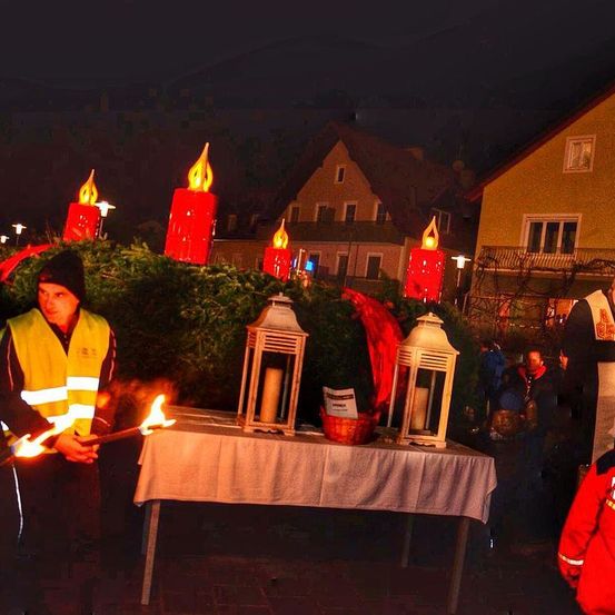 In einer Nachtszene stehen mehrere Personen um einen Tisch mit brennenden Kerzen. Eine Person hält brennende Fackeln, und andere sind in Warnwesten. Gebäude und Grünpflanzen sind im Hintergrund.