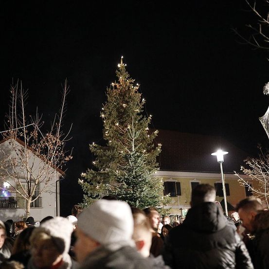 Ein großer beleuchteter Weihnachtsbaum steht bei Nacht, umgeben von Menschen. Der Baum ist mit Lichtern geschmückt, und eine Straßenlaterne beleuchtet das Gebiet. Ein Gebäude mit Balkon ist im Hintergrund sichtbar.