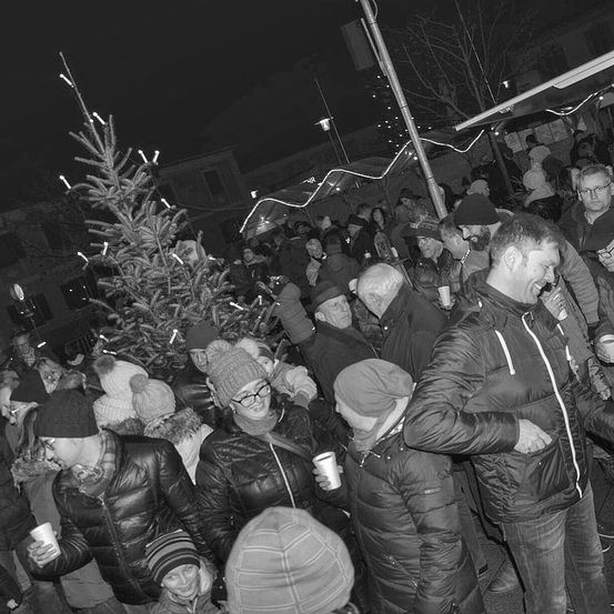Eine Menschenmenge, einige mit Wintermützen und Brillen, versammelt sich bei Nacht um einen beleuchteten Weihnachtsbaum mit Kerzen.