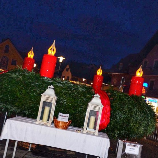 Ein festlicher Tisch mit roten Kerzenlaternen steht unter einem Nachthimmel, umgeben von immergrünen Pflanzen. Zwei brennende Kerzen in Laternen und ein kleiner Korb stehen auf dem Tisch.