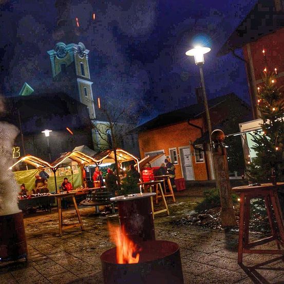 Eine Nachtmarktszene mit beleuchteten Ständen, einem Weihnachtsbaum und einem Turm im Hintergrund. Menschen versammeln sich um Tische, und ein Feuerkorb brennt hell.