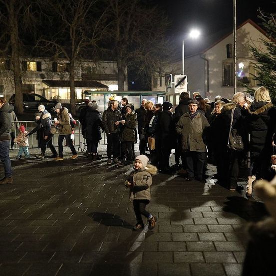Eine Gruppe von Menschen versammelt sich nachts auf einem Stadtplatz. Ein Kind läuft vor ihnen. Einige Leute stehen in der Nähe der Absperrung. Eine Straßenlaterne ist rechts.