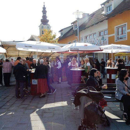 Eine geschäftige Marktszene mit Menschen unter Schirmen. Verkäufer und Käufer sind anwesend, ein Glockenturm ist im Hintergrund sichtbar.