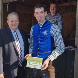 Ein Mann in einer blauen Weste steht zwischen zwei Männern, einer in einem Anzug, und hält ein Zertifikat mit '1. Platz' in den Händen. Sie stehen vor einer Holzwand.