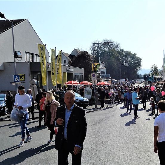 Eine belebte Straße mit vielen Fußgängern. Einige tragen Taschen, andere halten Luftballons. Es gibt Gebäude mit Bannern an der Seite und geparkte Autos in der Nähe.