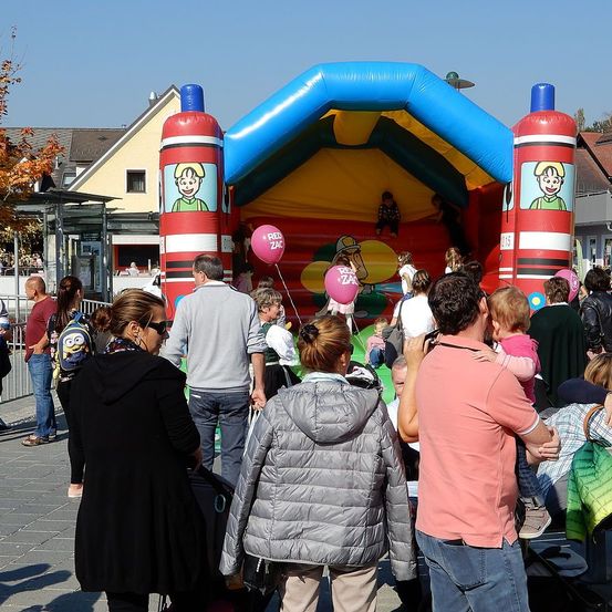 Eine Gruppe von Menschen, einschließlich Kindern, versammelt sich um ein aufblasbares Hüpfburg in einem öffentlichen Raum. Die Hüpfburg zeigt Zeichentrickfiguren.