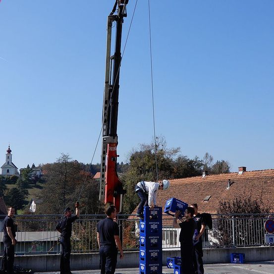 Eine Gruppe von Arbeitern stapelt Kisten auf einer Brücke unter einem klaren blauen Himmel. Ein Kran hebt eine Kiste, während andere den Stapel halten. Bäume und ein Haus sind im Hintergrund.