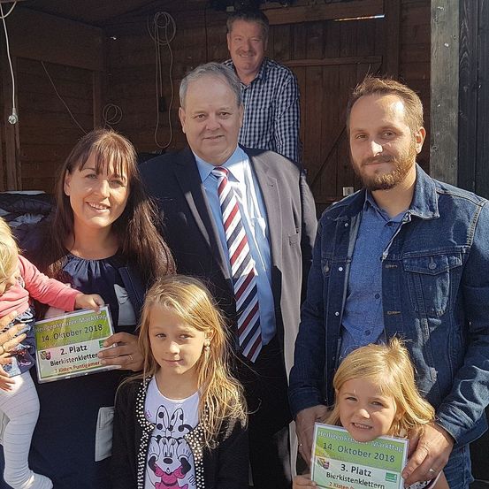 Eine Familie und zwei Männer posieren für ein Foto. Ein Mann hält einen Ausweis und ein anderer ein kleines Kind. Die Umgebung scheint ein Schuppen zu sein.