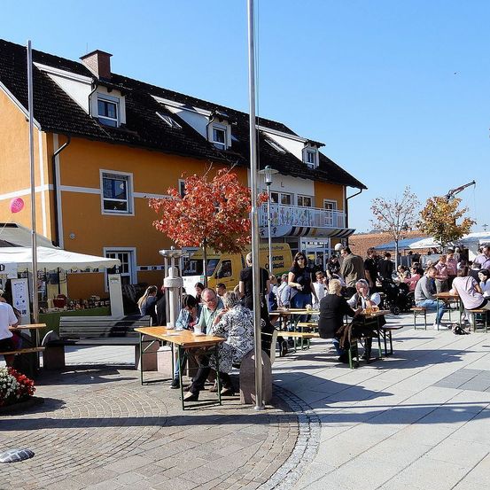 Ein gelbes Gebäude mit mehreren Fenstern im Hintergrund. Die Leute sitzen und stehen an den Tischen draußen. Das Wetter ist sonnig.