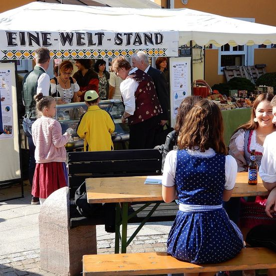 Eine Gruppe von Menschen in traditioneller bayerischer Kleidung versammelt sich um einen Imbissstand. Ein kleines Mädchen in einem rosa Kleid beobachtet, wie das Essen zubereitet wird.
