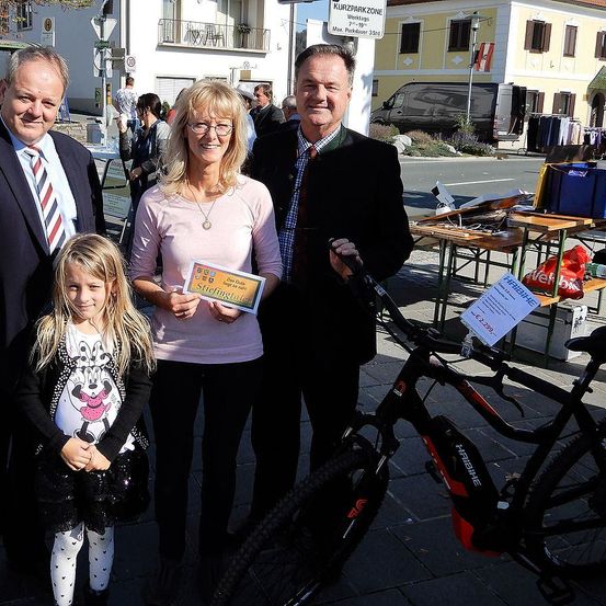 Drei Erwachsene und ein Kind stehen vor einem Fahrrad und lächeln für ein Foto. Sie halten ein Zertifikat in der Hand und sind von einer belebten Straße mit geparkten Autos, Tischen und einem Gebäude mit einem Schild umgeben.