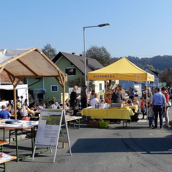 Ein gelbes Zelt-Markt ist in einem Außenbereich aufgestellt, wo sich viele Menschen versammelt haben. Eine Straßenlaterne steht am Straßenrand. Ein Tisch mit gelber Tischdecke hat viele Gegenstände darauf. Ein Schild steht vor dem Tisch.