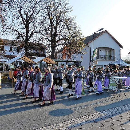 Eine Musikkapelle aus Männern und Frauen in traditionellen Kostümen spielt ihre Instrumente bei einem Outdoor-Umzug vor Häusern und Bäumen.