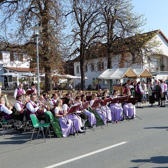Eine Straßenband spielt auf einem Stadtplatz, mit Musikern in lila Kostümen, die auf grünen Stühlen sitzen. Zuschauer und Zelte sind in der Nähe.
