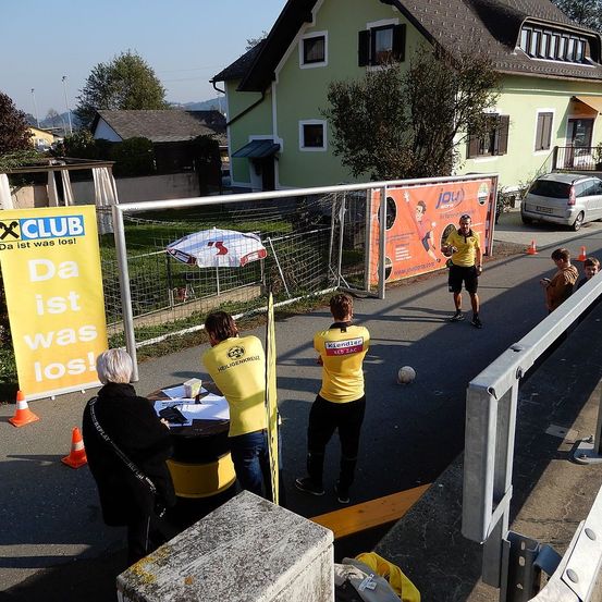 Ein Outdoor-Fußballevent findet statt. Menschen stehen und gehen in der Nähe eines Tisches mit Papieren und einem Becher. Ein Fußballtor ist aufgestellt und ein Ball ist in der Nähe.