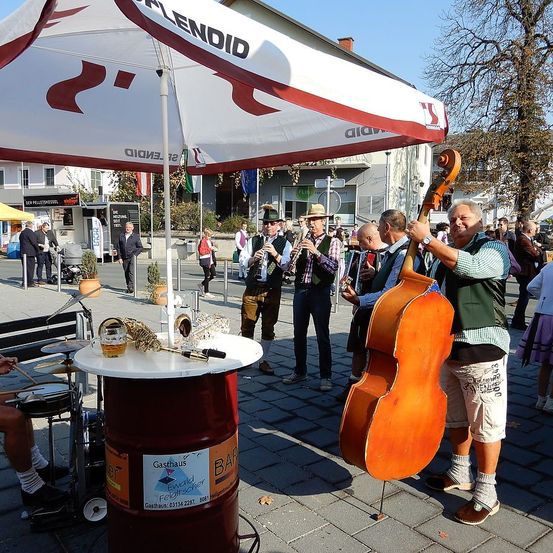 Eine Band spielt Musik auf einer Stadtstraße. Ein Mann hält ein Cello, während ein anderer Akkordeon spielt. Ein Fass dient als improvisiertes Schlagzeug.