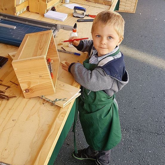 Ein junger Junge in einer grünen Schürze steht an einem Holzarbeitstisch und hält einen roten Stift in der Hand und lächelt. Hinter ihm befinden sich verschiedene Werkzeuge und Materialien.