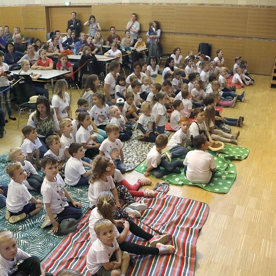 Eine große Gruppe von Kindern und Erwachsenen versammelt sich in einem Raum, wahrscheinlich für eine Veranstaltung. Die Kinder sitzen auf Decken, während einige stehen. Sie tragen weiße T-Shirts mit verschiedenen Designs. Einige von ihnen halten etwas, möglicherweise ein Musikinstrument. Der Raum hat Holzboden und Wände.