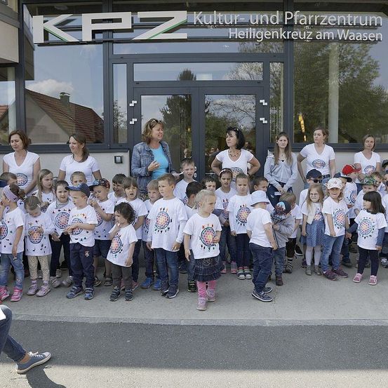 Eine Gruppe von Kindern und Erwachsenen steht vor dem Gebäude des Kultur-und Pfarrzentrums Heiligenkreuz am Waasen. Die Kinder tragen weiße T-Shirts mit einem kreisförmigen Design. Einige Erwachsene tragen Hüte und Schuhe. Das Gebäude hat Glastüren und Fenster.