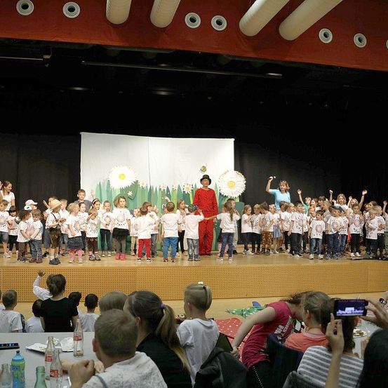 Eine Gruppe von Kindern in weißen T-Shirts tritt auf einer Bühne auf, mit einer Person in einem roten Kostüm und zwei Erwachsenen, die vorne stehen. Dahinter ist eine weiße Kulisse mit Blumen zu sehen. Das Publikum sitzt unterhalb der Bühne, einige machen Fotos.