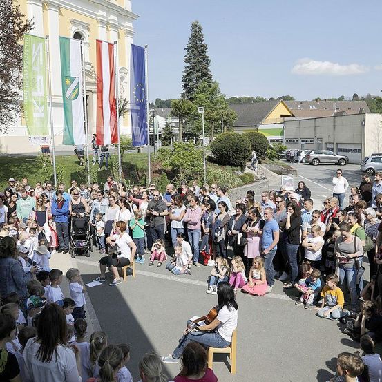 Eine Menschenmenge versammelt sich im Freien und beobachtet eine Frau, die vor einem Gebäude mit Fahnen Cello spielt. Bäume und ein geparktes Auto sind im Hintergrund zu sehen.