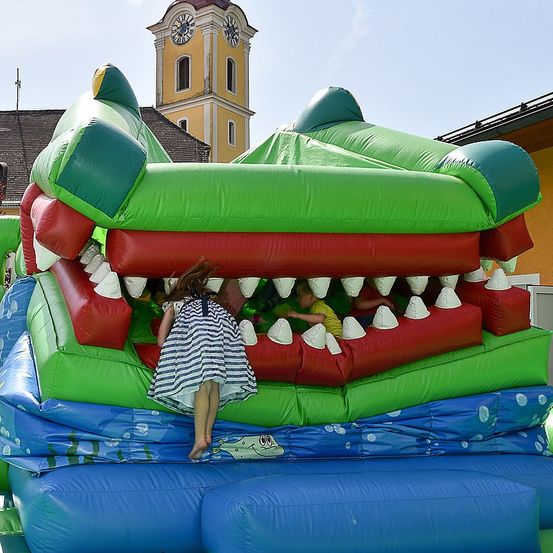 Kinder spielen in einem großen aufblasbaren Krokodil nahe eines Kirchturms. Das aufblasbare Objekt ist rot und grün.