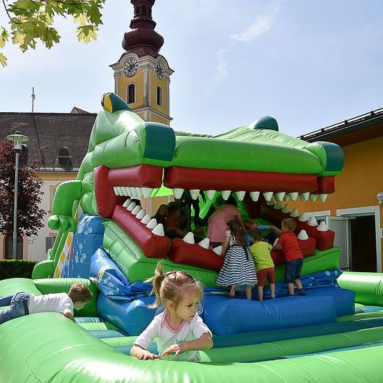 Kinder spielen in einem großen aufblasbaren Krokodil in einem Park mit einem Uhrturm im Hintergrund.