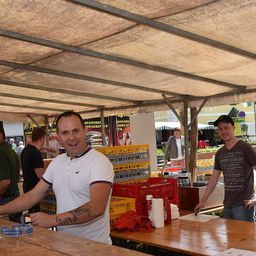 Zwei Männer lächeln unter einem Baldachin auf einem Markt. Einer hat eine Tätowierung am Arm und hält einen blauen Behälter. Der andere schneidet Gemüse auf einem Holztisch.