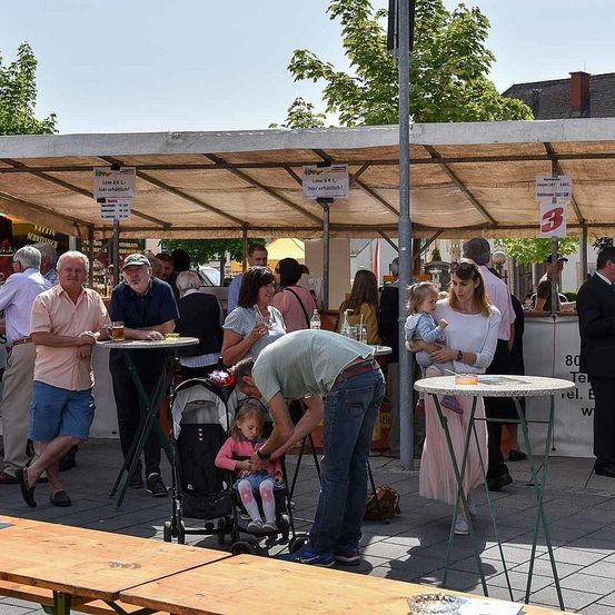 Eine Gruppe von Menschen steht unter einem Vordach auf einem Markt im Freien. Ein Mann beugt sich vor, um einem Kind in einem Kinderwagen zu helfen. Es gibt Tische mit Getränken und Essen.