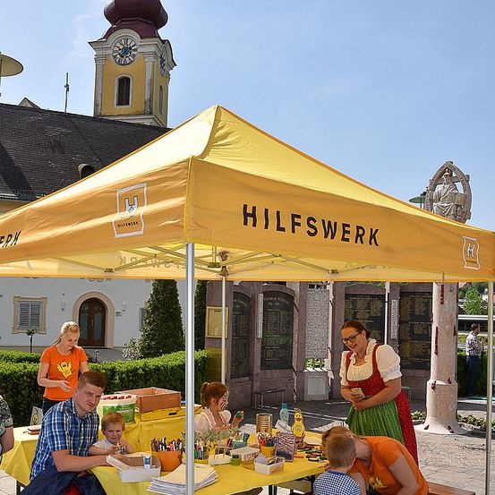 Eine Gruppe von Menschen sitzt unter einem gelben Zelt mit der Aufschrift 'Hilfswerk'. Sie sind mit einer Aktivität beschäftigt und haben Kunstmaterial auf dem Tisch. Im Hintergrund steht eine Kirche mit einem Turm und einer Uhr.