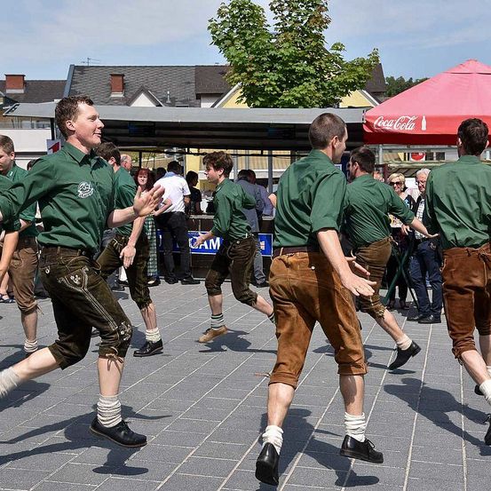Eine Gruppe von Männern in traditioneller deutscher Kleidung, möglicherweise an einer Parade teilnehmend, tanzt auf einem Kopfsteinpflasterplatz unter einem roten Regenschirm. Dahinter befindet sich ein Stand mit einem Coca-Cola-Schild und Zuschauern.