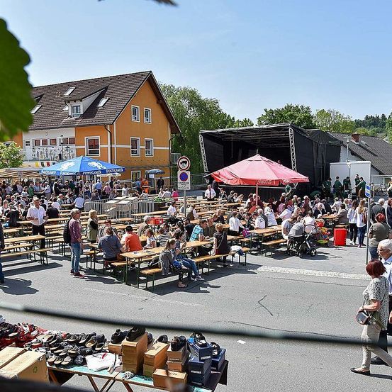 Ein überfüllter Außenmarkt mit vielen Menschen, die unter Schirmen an Tischen sitzen. In der Nähe befindet sich eine Bühne mit einem laufenden Event und einem Gebäude mit Schildern. Im Vordergrund werden Schuhe auf einem Tisch ausgestellt.