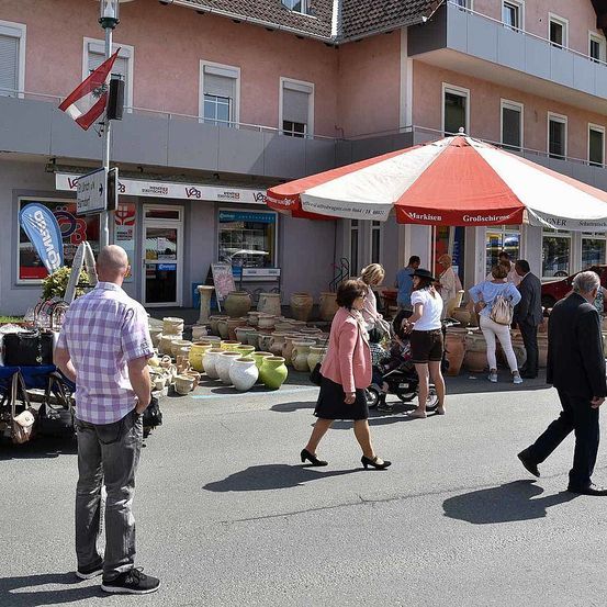 Menschen schlendern auf einer belebten Straße mit verschiedenen Geschäften und Außengastronomie unter einem Regenschirm. Ein Mann steht neben einem Warenwagen, während andere stöbern und sich unterhalten.