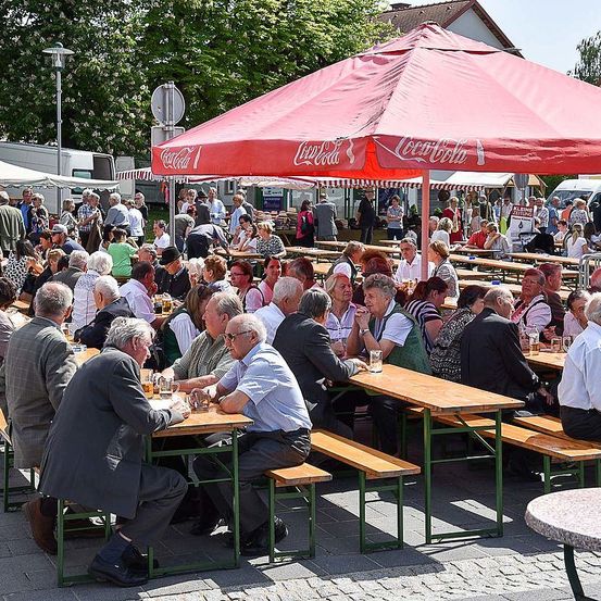 Eine Gruppe von Menschen sitzt unter einem roten Coca-Cola-Regenschirm an Tischen, wahrscheinlich in einem Biergarten. Einige trinken Bier, während andere in Gespräche vertieft sind.