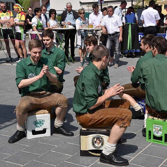 Eine Gruppe von Männern in grünen Hemden und braunen Hosen sitzt auf Kisten und klatscht. Hinter ihnen stehen und sitzen Leute um Tische herum. Einige tragen Radtrikots, andere haben Freizeitkleidung an. Im Hintergrund sind Gebäude und Grün zu sehen.